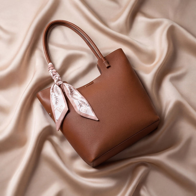 A structured brown bag with dual top handles, adorned with a pink and white patterned silk scarf tied to the handle